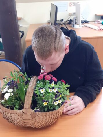 Na zdjęciu chłopak siedzi przy stole. Chłopak wącha kwiaty wiosenne, które są umieszczone w koszyczku. W koszyczku są bratki, stokrotki i hiacynt.