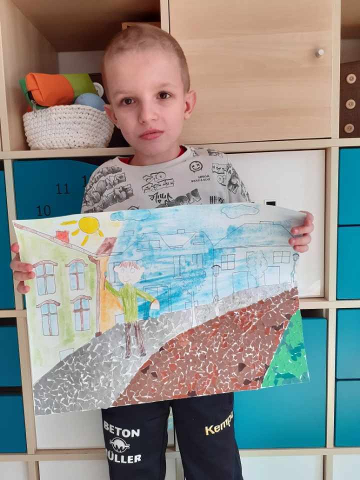 Na zdjęciu stoi chłopiec trzyma pracę plastyczna na której wyklejona jest droga i chodnik skrawkami papieru, wzdłuż kamienice jedna brązowa reszta niebieska oraz idący człowiek.