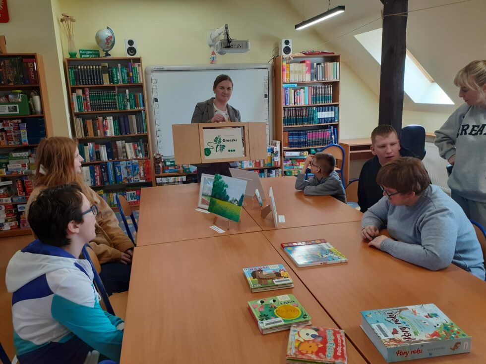 Na zdjęciu chłopcy i dziewczynki siedzą przy stole na krzesłach. Na stole leżą kolorowe książki. Przed stołem stoi Pani bibliotekarka, trzyma drewniany teatrzyk w którym jest bajka o groszku. W tle regały z książkami. 