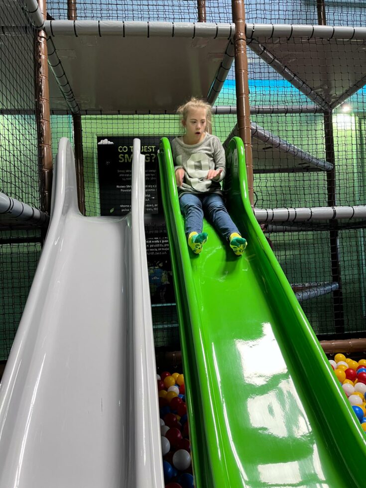 Na zdjęciu dziewczynka zjeżdża po zielonej zjeżdżalni. W tle platformy i siatki w dole kolorowe, plastikowe piłki.