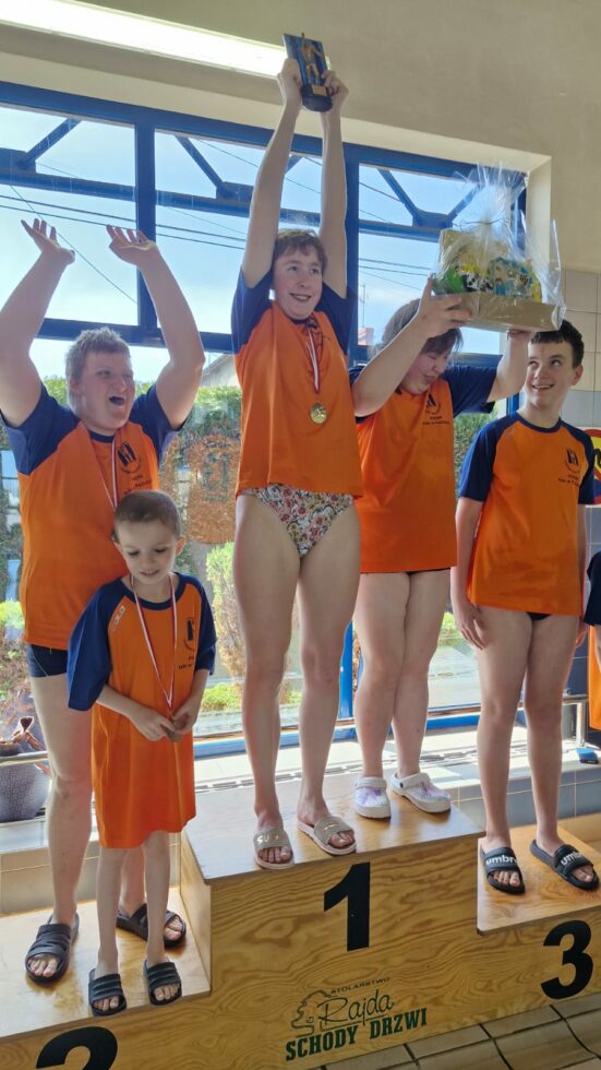 Na zdjęciu stoją chłopcy i dziewczyny w pomarańczowo granatowych koszulkach na stopniach podium. Na szyi maja medale. Jedna dziewczyna unosi do góry puchar druga upominki.