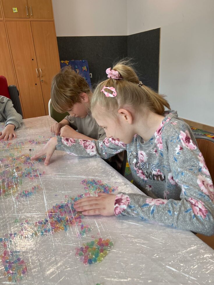 Na zdjęciu przy stole siedzi dziewczynka i chłopiec. Na stole leżą kolorowe kulki hydrożelowe przykryte folią. Dzieci paluszkami przesuwają kolorowe kulki hydrożelowe.