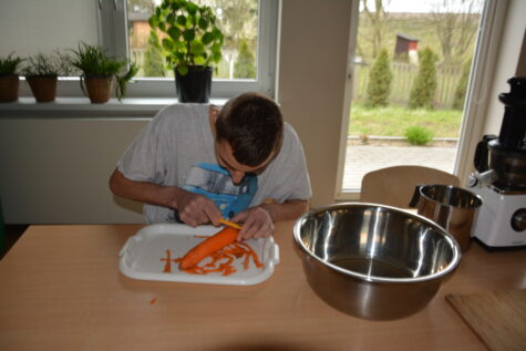 Na zdjęciu siedzi chłopak przy stole na białej tacce ma marchewkę którą obiera. Na stole stoi blaszana miska i wyciskarka.