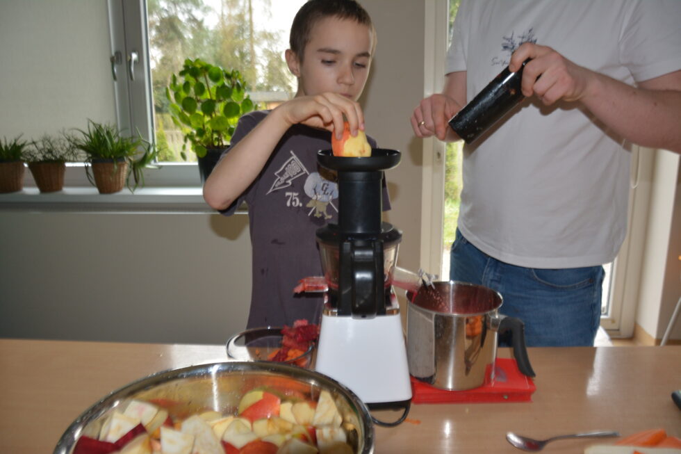 Na zdjęciu chłopiec stoi przy stole i wkłada kawałek jabłka do wyciskarki, obok stoi opiekun. Na stole stoi miska z obranymi warzywami, jabłka , marchew. 