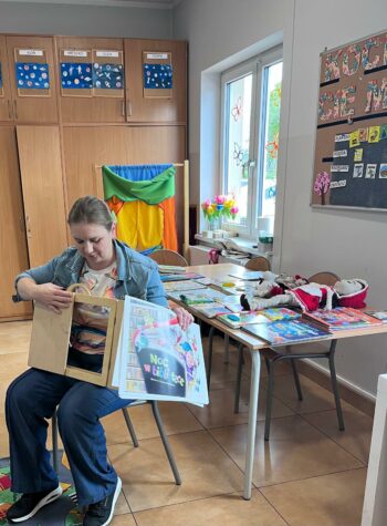 Na zdjęciu kobieta siedzi na krześle, trzyma drewniany teatrzyk z bajką. W tle stół i szafa oraz okno. Zdjęcie w sali lekcyjnej.