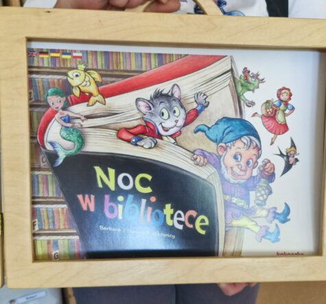 Na zdjęciu drewniany teatrzyk i pierwsza strona z tytułem bajki Noc w bibliotece. Obrazek przedstawia książkę z której stronic wychodzą postaci z bajek, krasnoludek, kot w butach, czerwony kapturek, złota rybka, mała syrenka, smok i calineczka.