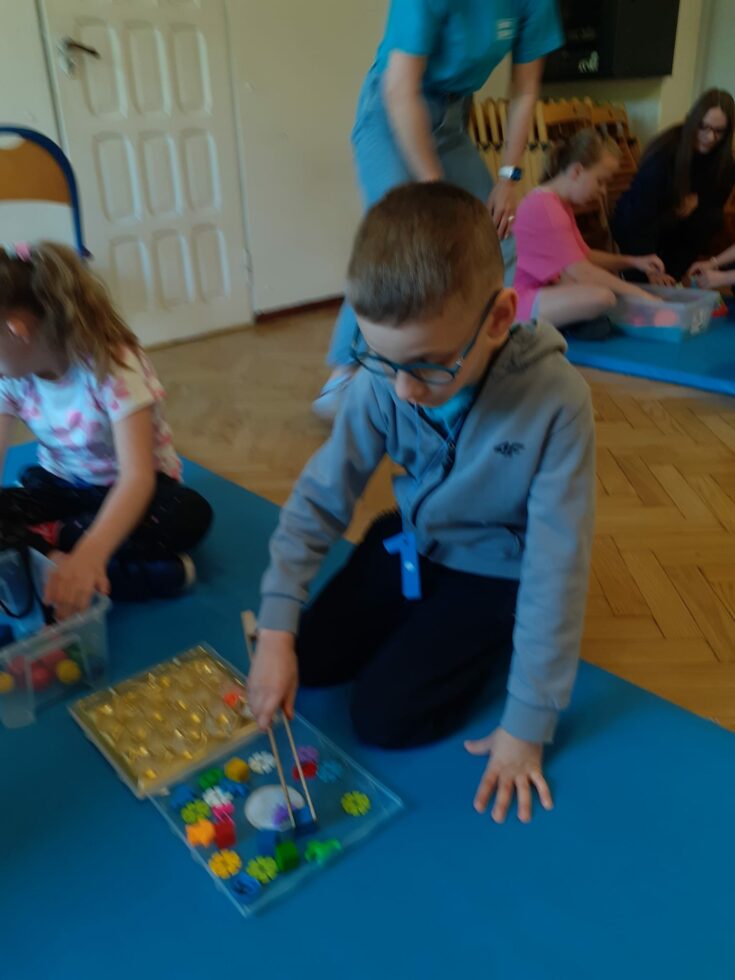 Na zdjęciu chłopiec w okularach klęczy na niebieskim materacu. W prawej ręce ma drewniane szczypce. Przenosi szczypcami kolorowe plastikowe kwiatki do pojemnika z wgłębieniami.