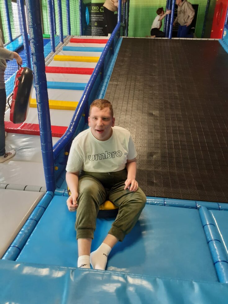 Na zdjęciu chłopak siedzi na dole dużej zjeżdżalni w sali zabaw. W tle inne dzieci.
