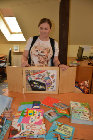 Na zdjęciu stoi kobieta przy stole na którym leżą kolorowe książki. Kobieta trzyma drewniany teatrzyk w którym jest tytułowa ilustracja opowiadania "Noc w bibliotece". W tle biurko i okno dachowe.