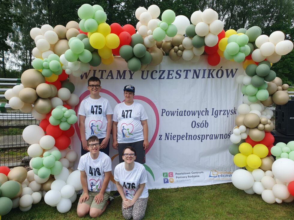 Na zdjęciu baner z napisem witamy uczestników powiatowych igrzysk osób z niepełnosprawnością. Dookoła banera przypięte kolorowe balony. Przed banerem czterech zawodników jedna dziewczyna i trzech chłopców.
