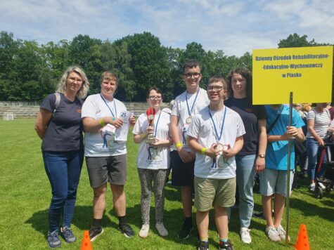 Na zdjęciu zawodnicy w koszulkach z numerami i medalami na szyi. Jedna dziewczyna i trzech chłopców oraz dwie opiekunki. obok stoi chłopak z żółta tablica z napisem ośrodka.
