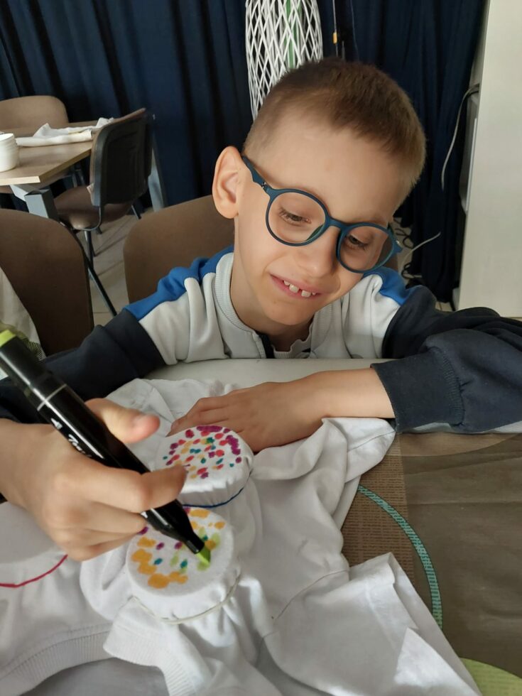 Na zdjęciu chłopiec w okularach siedzi przy stole na krześle. W prawej ręce trzyma zielony mazak i robi kropki na białej tkaninie. Tkanina naciągnięta jest na słoik i przytrzymana gumką.