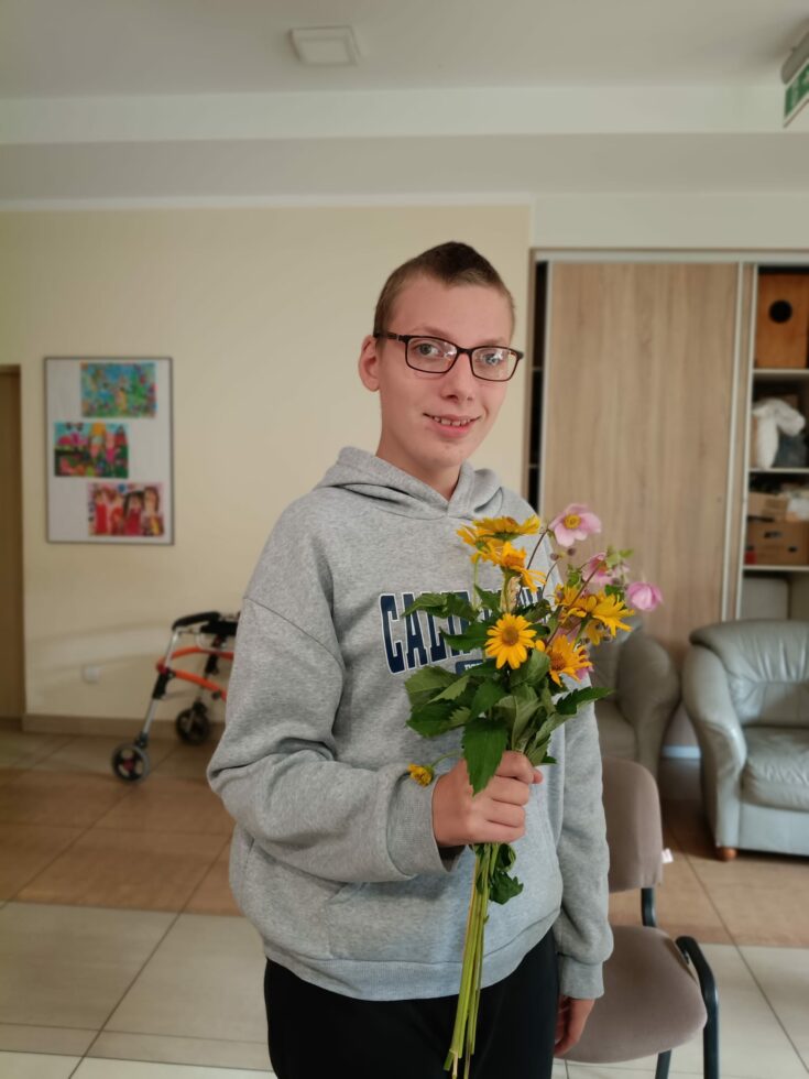Na zdjęciu stoi chłopak w okularach, w prawej ręce trzyma bukiet z żółtych i różowych kwiatów.