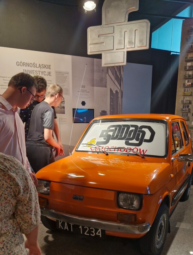 Na zdjęciu pomarańczowy Fiat 126 P obok stoją trzej chłopcy w okularach i oglądają auto.