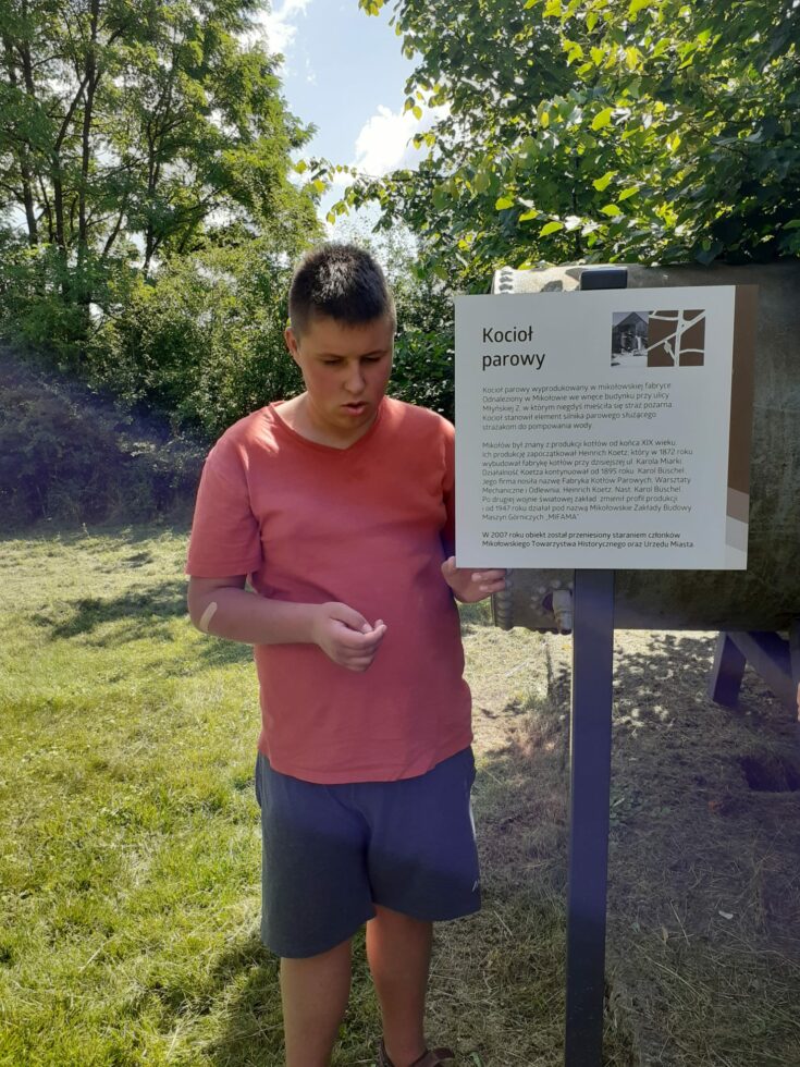 Na zdjęciu stoi chłopak w czerwonej koszulce i granatowych spodenkach. Obok tablica informacyjna z napisem kocioł parowy. W tle drzewa i krzewy.