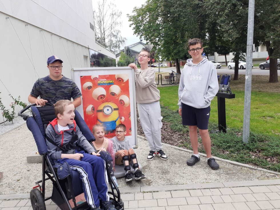 Na zdjęciu stoją trzej chłopcy przy plakacie filmu o Minionkach, dziewczynka z chłopcem siedzą przed plakatem, czwarty chłopiec siedzi w wózku terapeutycznym. W tle budynek im drzewa.