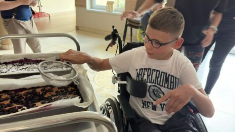 Na zdjęciu siedzi chłopiec w wózku terapeutycznym w okularach. W prawej ręce trzyma sitko z cukrem pudrem. Chłopiec posypuje ciastom które stoi na wózku.
