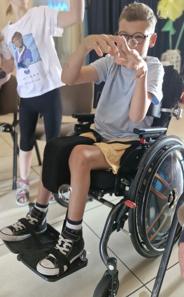 Na zdjęciu chłopiec w okularach siedzi na wózku inwalidzkim, w tle inne dziecko. Chłopiec unosi ręce do góry próbując dotknąć bańkę mydlaną.