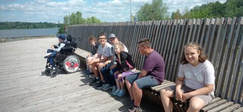 Na zdjęciu na ławce usytuowanej na długim pomoście siedzą chłopcy i dziewczynki. Jeden chłopiec siedzi na specjalistycznym wózku inwalidzkim. W tle jezioro i drzewa.