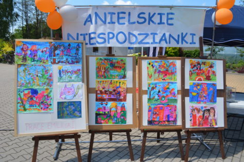 Na zdjęciu cztery sztalugi z kolorowymi pracami plastycznymi w tle napis Anielskie Niespodzianki, zdjęcie z festynu.