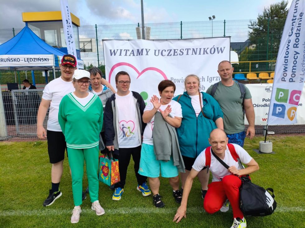 Na zdjęciu uczestnicy Świetlicy Terapeutycznej wraz z opiekunem stoją na murawie boiska w tle banery przed igrzyskami sportowymi.