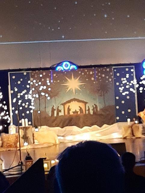 Na zdjęciu scena udekorowana świecami, w tle podświetlana szopka. 