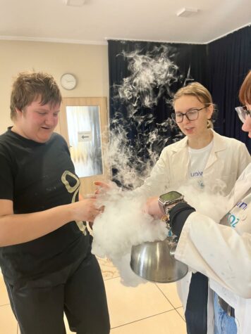 Na zdjęciu dwie kobiety w okularach trzymają garnek z którego unosi się biała mgła obok stoi chłopak.