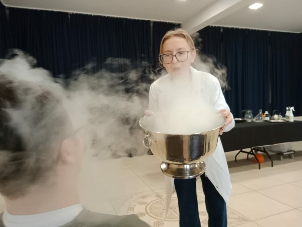 Na zdjęciu stoi kobieta w okularach w białym fartuchu. W rękach trzyma metalowe naczynie z którego unosi się biały dym. Stoi na przeciw mężczyzny pokazując mu eksperyment.