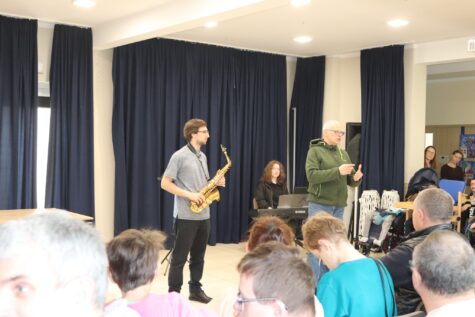 Na zdjęciu mężczyzna z saksofonem, kobieta przy klawiszach i mężczyzna w okularach oraz widzowie.