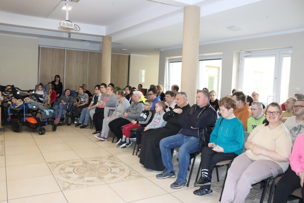 Na zdjęciu mężczyźni, kobiety, chłopcy, dziewczynki, ksiądz siedzą na krzesłach podczas warsztatów muzycznych.