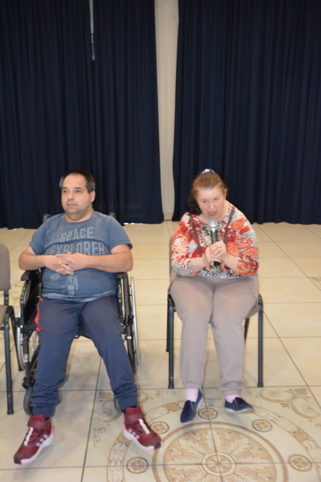 Na zdjęciu siedzi mężczyzna na wózku i kobieta siedzi na krześle. Kobieta trzyma mikrofon.