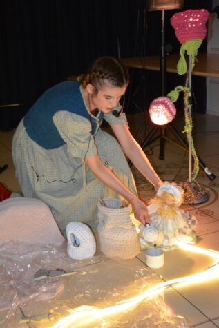 Na zdjęciu kobieta kuca, w lewej ręce trzyma lalkę, prawą czajniczkiem nalewa wodę do filiżanki. Obok podświetlony kwiatek z włóczki.