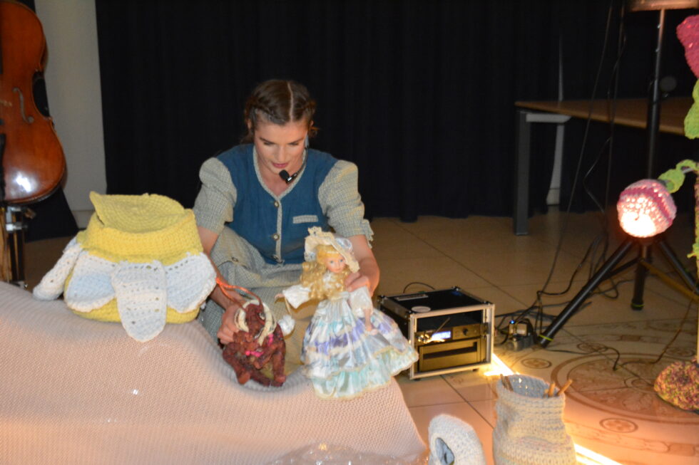 Na zdjęciu kobieta kuca. W prawej ręce trzyma dużą kukiełkę pszczoły w lewej ręce lalkę w sukni i kapeluszu. Obok stoi zrobiony na szydełku kwiat stokrotki żółto biały.