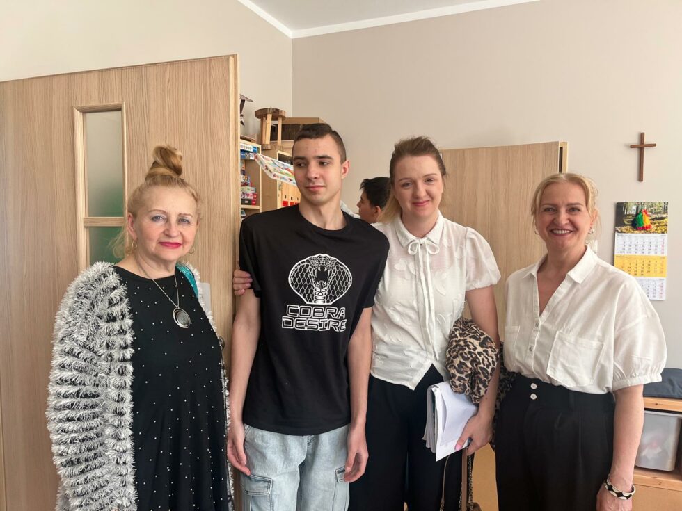 Na zdjęciu trzy kobiety w różnym wieku stoją z nastoletnim chłopakiem. W tle regał z puzzlami i chłopak wkładający na półkę kolorowe pudełko.