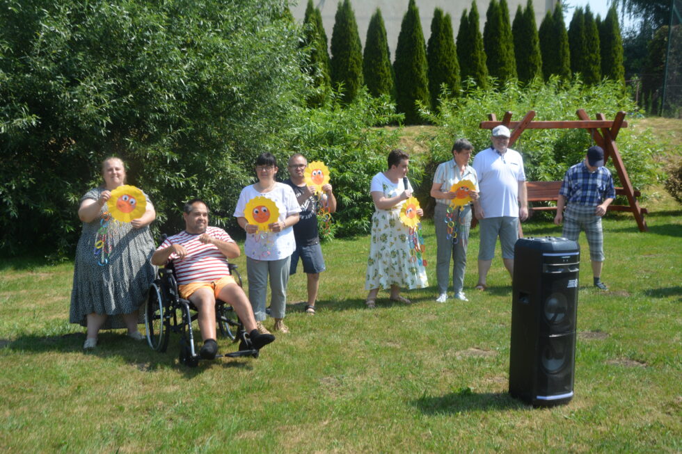 Na zdjęciu kobiety i mężczyźni uczestnicy Świetlicy Terapeutycznej, stoją na trawie, jeden mężczyzna siedzi na wózku inwalidzkim. W rękach trzymają żółte słoneczka wykonane z papieru. Jedna z kobiet trzyma w prawej ręce mikrofon. Przed nimi na trawie stoi kolumna, w tle zielone krzewy. 