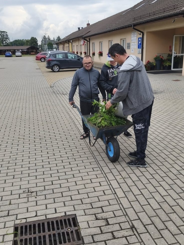 Na zdjęciu trzej chłopcy , jeden trzyma taczkę, dwóch wrzuca do niej chwasty. W tle budynek i zaparkowane auta.