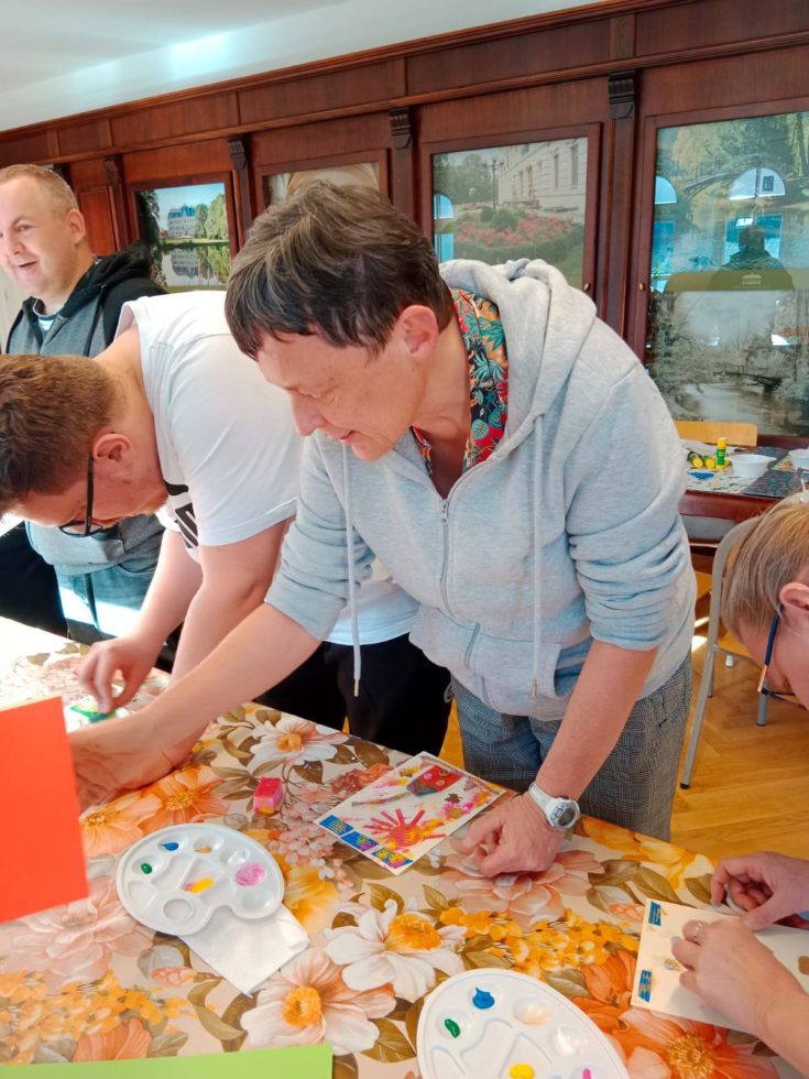 Na zdjęciu kobieta i dwaj mężczyźni stoją przy stole wykonując prace plastyczne podczas warsztatów z kolagrafii kolorowymi farbami.