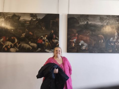 Na zdjęciu stoi kobieta w tle dwa obraz w Muzeum Zamkowym w Pszczynie przedstawiające czynności ludzi mieszkających na wsi.