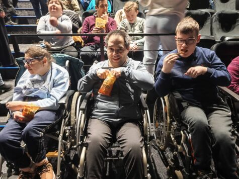 Na zdjęciu kobieta i dwóch mężczyzn na wózkach inwalidzkich w sali kinowej.