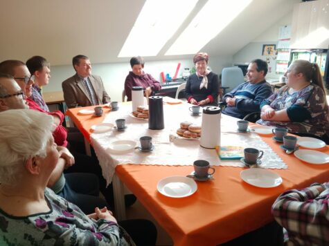 Na zdjęciu siedzą przy stole kobiety i mężczyźni uczestnicy ?Świetlicy Terapeutycznej podczas spotkania z księdzem. na stole stoją termosy, filiżnki oraz talerzyki. Na środku stołu stoją dwa talerze z pączkami.