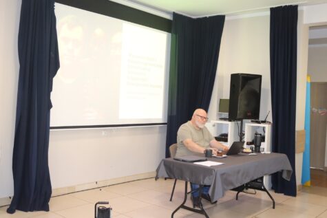 Na zdjęciu siedzi mężczyzna na krześle przy stole, za nim ekran projektora. Zdjęcie podczas szkolenia.