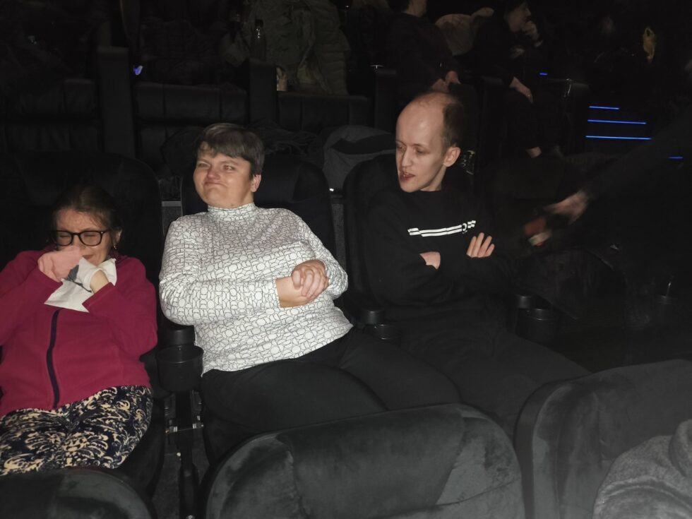 Na zdjęciu dwie kobiety i mężczyzna, uczestnicy Świetlicy Terapeutycznej siedzą na czarnych fotelach w kinie w oczekiwaniu na seans filmowy. , 