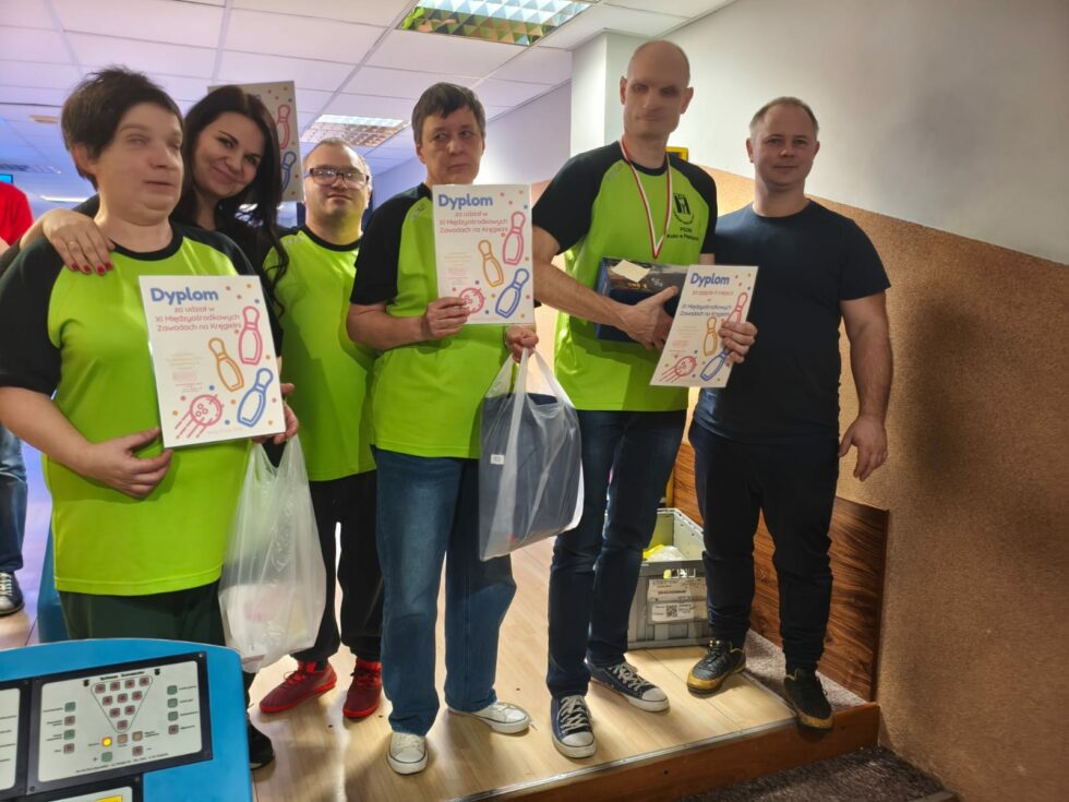 Na zdjęciu mężczyźni i kobiety uczestnicy Świetlicy Terapeutycznej w zielono granatowych koszulkach wraz z opiekunami stoją z dyplomami po zawodach w kregla.