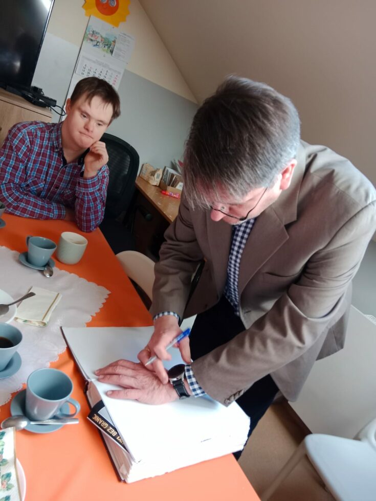 Na zdjęciu mężczyzna pochylony nad biała kartką w grubej księdze. W prawej ręce trzyma długopis. Księga leży na stole obok filiżanki koloru szarego. Przy stole siedzi chłopak i patrzy.