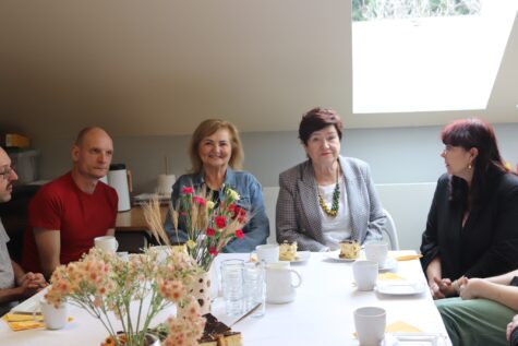 Na zdjęciu siedzą trzy kobiety przy stole, dwóch mężczyzn. Na stole stole stoja kwiaty w wazonie, białe filiżanki i talerzyki z sernikiem.