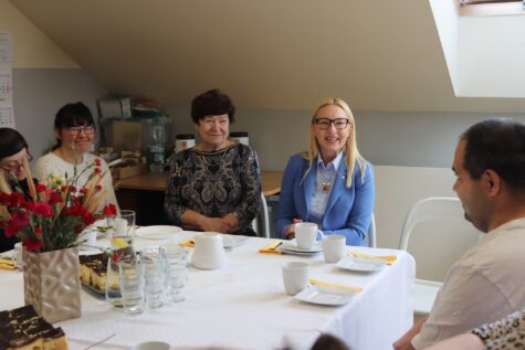 Na zdjęciu przy stole siedzą dwie kobiety, jedna ma okulary i długie blond włosy, druga jest szatynka z krótką fryzura. Przy stole siedzą również dwie kobiety i mężczyzna, uczestnicy Świetlicy Terapeutycznej. Na stole leży obrus i stoją filiżanki i talerzyki, czerwone goździki w wazonie i pokrojony sernik na prostokątnym talerzu.