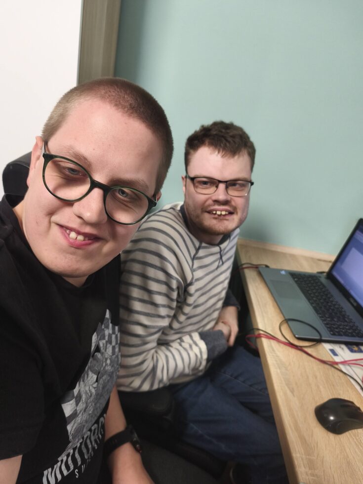 Na zdjęciu dwaj mężczyźni w okularach siedzą przy biurku, na którym stoi laptop.