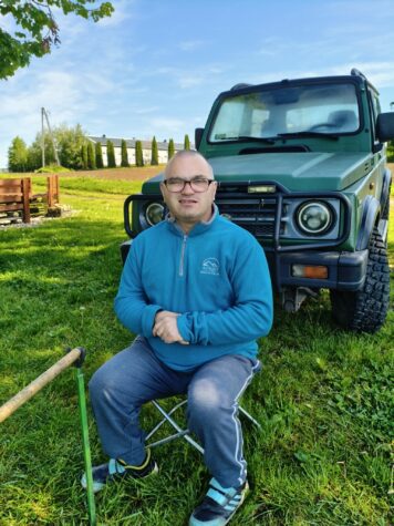 Na zdjęciu siedzi mężczyzna w okularach na krzesełku wędkarskim, przed nim stoi wędka. W tle zielony samochód.