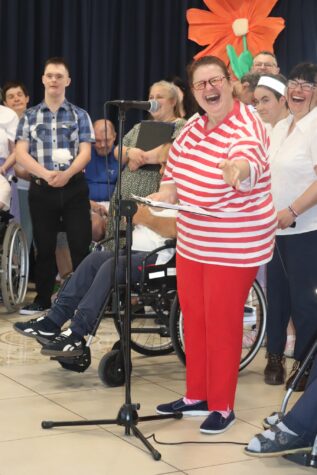 Na zdjęciu uśmiechnięta kobieta w czerwonych spodniach i koszulce w paski biało czerwone w okularach stoi przed mikrofonem. Na głowie ma czerwoną opaskę. W tle kobiety i mężczyźni. Podczas występu na Dzień Matki.