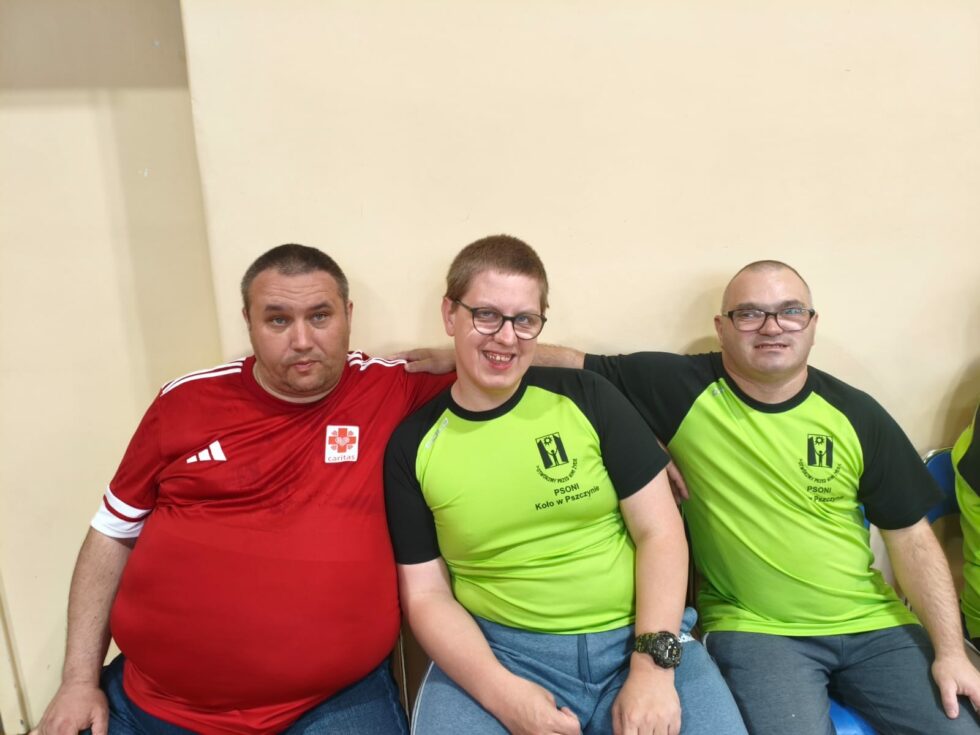 Na zdjęciu siedzą trzej mężczyźni. Jeden ma czerwoną sportową koszulkę z napisem i znaczkiem Caritas. Dwaj pozostali maja koszulki zielono czarne z napisem PSONI Koło w Pszczynie. Zdjęcie podczas zawodów w tenisa stołowego.